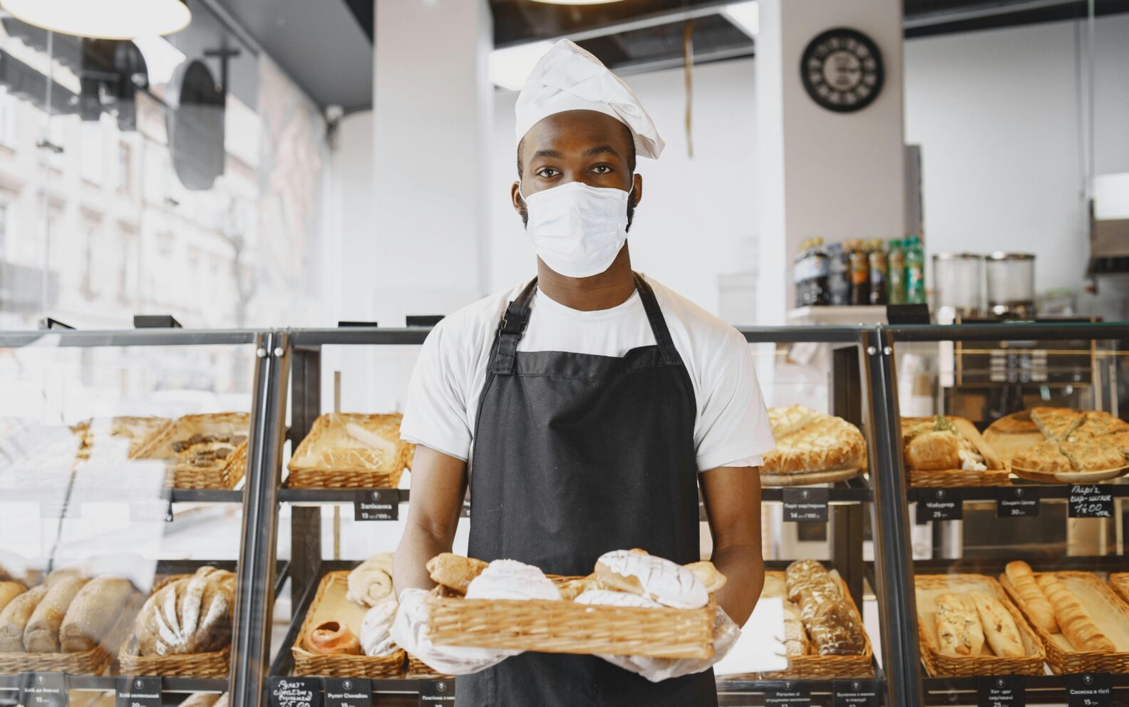 A baker wearing a face mask presents a tray of fresh bread in a modern bakery setting.